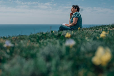 Adult woman thoughtfully writes plans diary on a sunny day on a mountain against backdrop of the sea