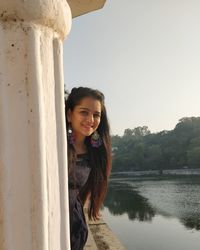 Portrait of smiling young woman standing by lake against sky