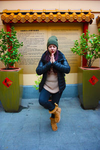 Smiling young woman praying while standing on one leg against wall