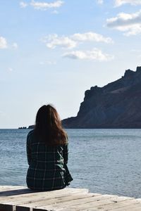 Rear view of woman sitting on rock by sea against sky