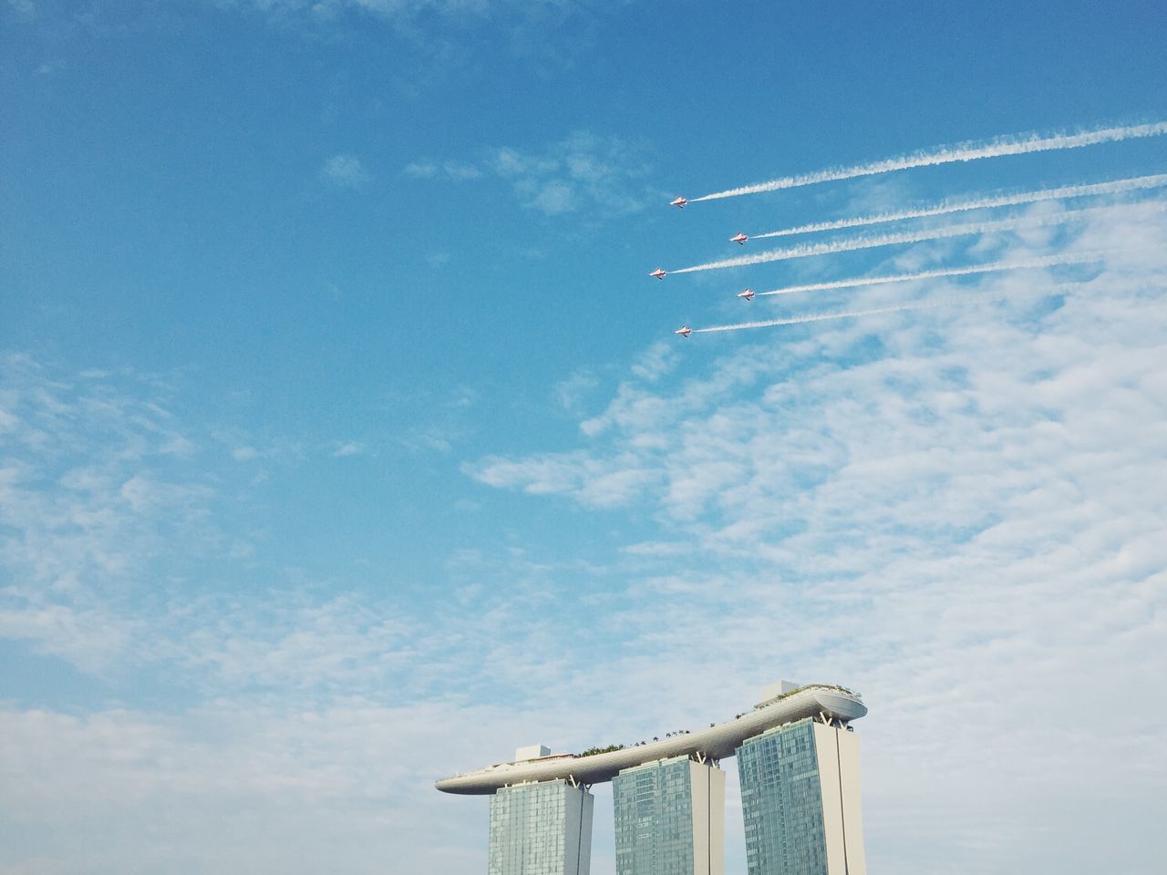 low angle view, sky, blue, airplane, built structure, cloud - sky, flying, air vehicle, transportation, architecture, travel, outdoors, day, mode of transport, no people, cloud, building exterior, flag, vapor trail, tall - high