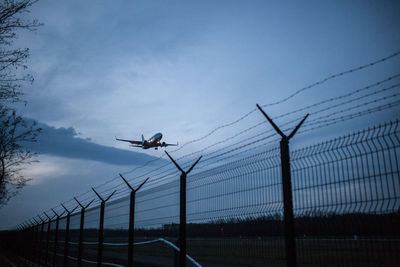 Low angle view of airplane flying over fence against sky at dusk