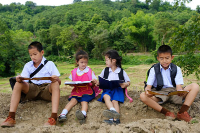 Siblings reading books while sitting on field against trees