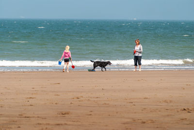 A young girl wears a pink floppy hat and carries a bucket and spade across a british beach.