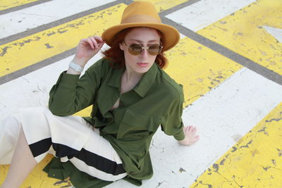 Portrait of ginger woman wearing green shirt and sunglasses with yellow felt hat sitting on road 