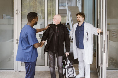 Senior male patient shaking hands with nurse while standing by doctor at hospital doorway