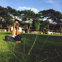 Woman photographing through camera while sitting on grassy field at park