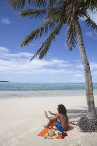 Male tourist with curly hair reading under palm tree at sandy beach