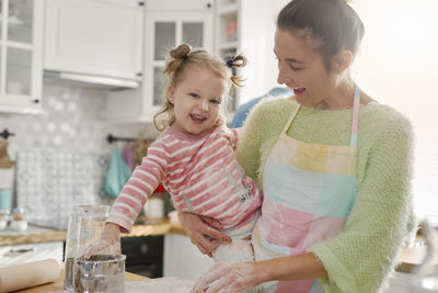 Mother carrying daughter while preparing food in kitchen at home