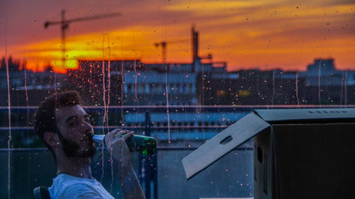 Young man drinking in balcony seen through wet window during sunset