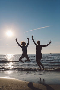 Cheerful female friends jumping at beach against sky on sunny day