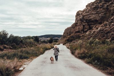 Rear view of woman walking with dog on road against sky