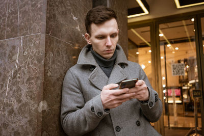 Smiling young man in gray coat is holding scroll text messages in his mobile