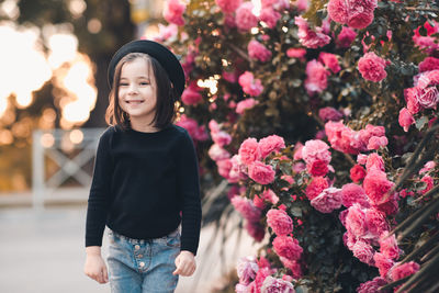 Cute smiling stylish child girl 5-6 year old wear hat and black top posing over rose flower outdoors