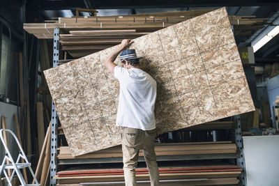 Rear view of man carrying wooden plank in front of shelf at workshop