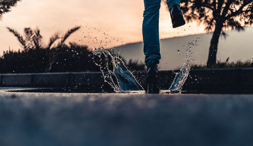 Low section of woman jumping on puddle against sky during sunset