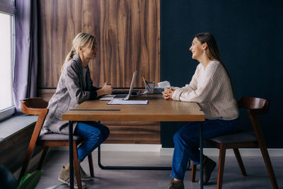 Two young modern business women smiling talking while sitting at the desk in the office.