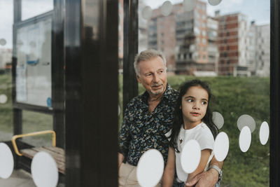 Senior man sitting with arm around granddaughter while sitting at bus stop