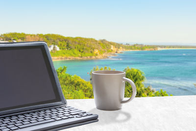 Coffee cup on table by sea against sky