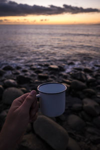 Close-up of hand holding cup against sea during sunset