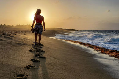 Full length rear view of woman standing on beach during sunset