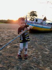 Cute boy holding rope of fishing boat while standing on shore against sky during sunset