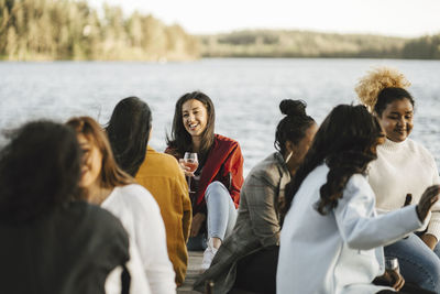 Smiling multiracial female friends sitting on jetty against lake