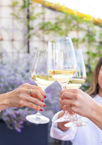 Cropped unrecognizable adult girlfriends in smart casual clothes sitting at restaurant table and clinking glasses of white wine during meeting on terrace