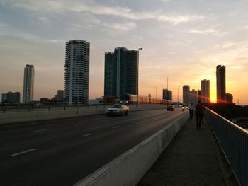 Road by buildings in city against sky during sunset