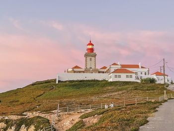 Lighthouse by building against sky during sunset