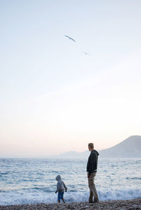 Rear view of man with son standing at beach against clear sky during sunset