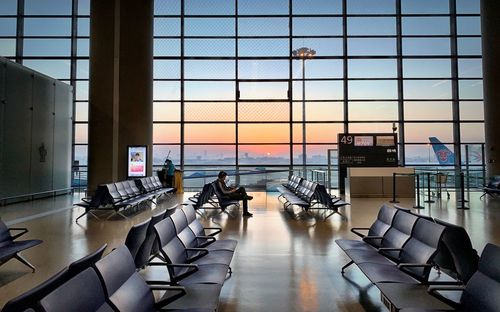 Empty chairs at airport against sky seen through glass window