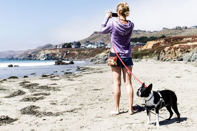 Full length of woman photographing on mobile phone with dog at beach