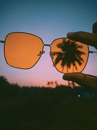 Hand holding sunglasses against sky during sunset