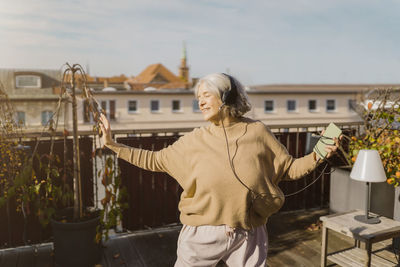 Carefree senior woman enjoying listening to music through headphones while standing at terrace