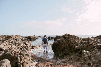 Rear view of young man standing at rocky shore against sky