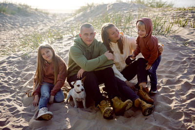 Family with a son and daughter and a dog sit on the sand in autumn