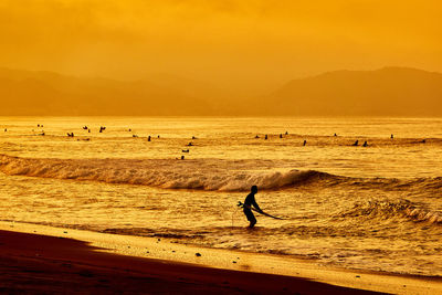 Scenic view of surfers beach with silhouette people against orange sky during sunrise