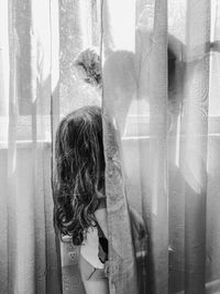 Rear view of young girl standing against curtain looking up at cat on windowsill