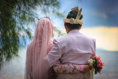 Rear view of couple standing at beach against cloudy sky during sunset