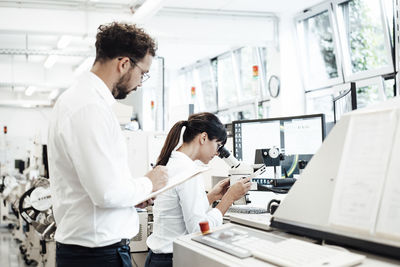 Male technician writing on clipboard while standing by female colleague looking through microscope at laboratory