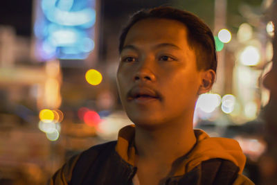Close-up of thoughtful young man looking away against illuminated lights at night