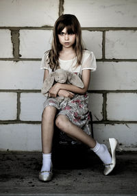 Full length portrait of girl with toy sitting against wall