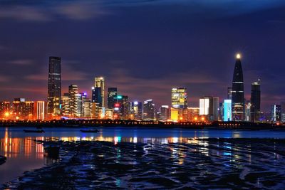 Illuminated buildings by river against sky at night