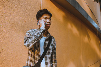 Young man talking on phone while standing by wall