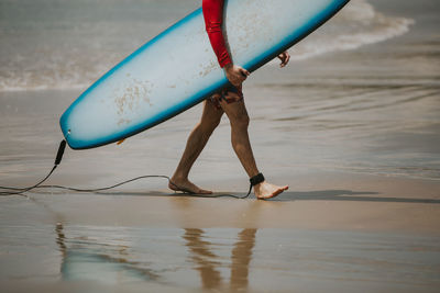 Low section of man carrying surfboard while walking at beach