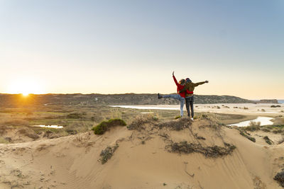 Young carefree couple with hand raised standing by man on sand dune during sunset