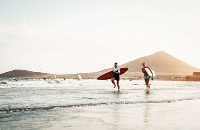 Man and woman running while holding surfboards at beach against sky