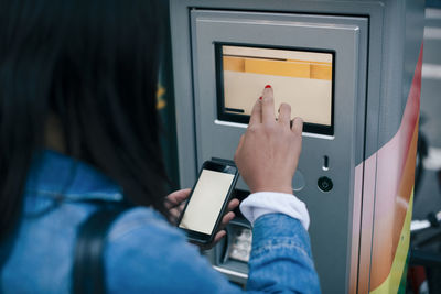 Rear view of teenage girl touching atm screen while holding smart phone