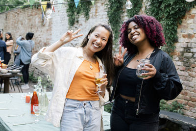 Portrait of happy female friends with drinks showing peace sign during dinner party in back yard